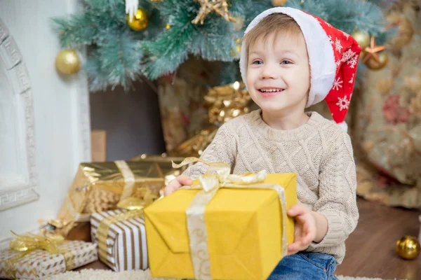 Kırmızı şapkalı bir çocuk bir Noel ağacı bir hediye ile oturuyor.