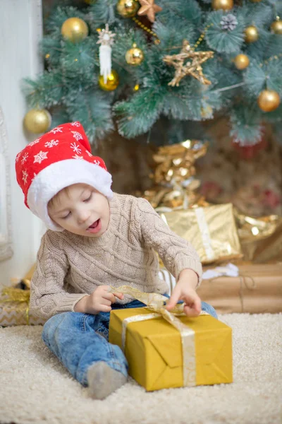 Kırmızı şapkalı bir çocuk bir Noel ağacı bir hediye ile oturuyor. Telifsiz Stok Fotoğraflar