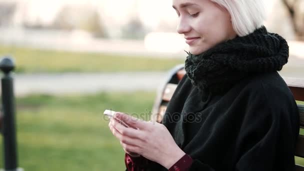 Pomp blond meisje buiten. Kijkt naar de smartphone zit op een houten bankje. Hij typt de tekst, glimlacht — Stockvideo