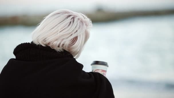 Menina loira na praia ele bebe café parece intodistance calmamente chamado ela olha para a câmera — Vídeo de Stock