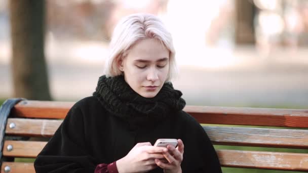 Blondes Mädchen blickt aufs Smartphone sitzt auf Bank bekommt Text in Smartphone ruhigen Gesichtsausdruck — Stockvideo