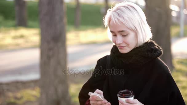 Blond dziewczyna na zewnątrz. W ręce smartphone i kawa. Typ tekst, uśmiechy, sprawia, że selfie — Wideo stockowe