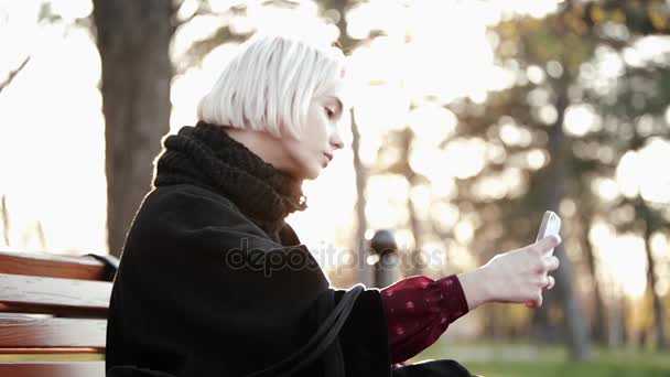 Blonde Mädchen draußen hält Smartphone und Kaffee machen ein Selfie zu einem ernsten schönen Gesicht Sonne blinkt — Stockvideo