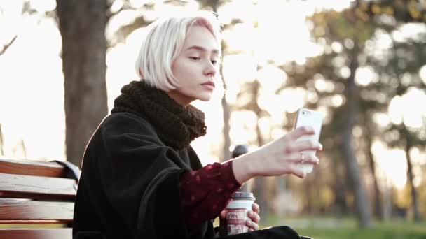 外のブロンドの女の子を保持するスマート フォンとコーヒーを作る、selfie 太陽点滅深刻な美しい顔 — ストック動画