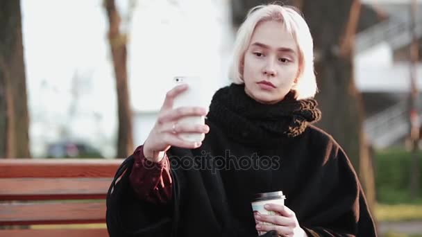 Ragazza bionda fuori tiene uno smartphone e caffè fanno un selfie un bel viso serio — Video Stock