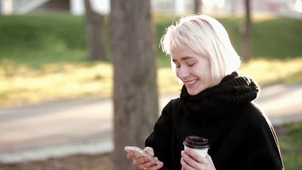 Draußen sitzt ein blondes Mädchen auf einer Holzbank und wählt den Text im Smartphone, lächelt und trinkt Kaffee. — Stockvideo