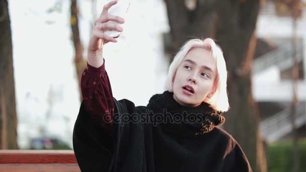 Ξανθιά κοπέλα εκτός κατέχει ένα smartphone και καφέ κάνουν μια selfie ένα σοβαρό πρόσωπο όμορφο ήλιο αναβοσβήνει — Αρχείο Βίντεο