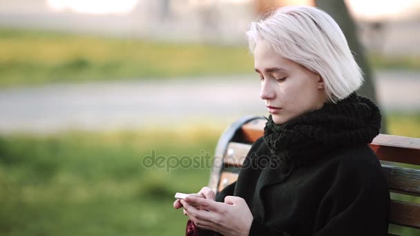 Chica rubia afuera Mira el teléfono inteligente en un banco Juega un juego activo en 3D en el teléfono inteligente Sonrisas — Vídeos de Stock