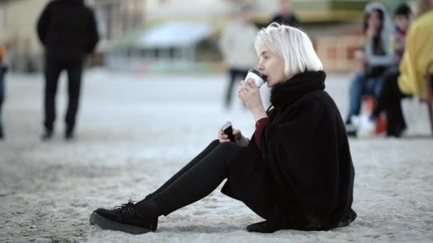 밖에 서 금발 소녀입니다. 해변에서 그가 커피를 마신다. 하얀 모래에 앉아 바 닷 바람을 즐긴다. — 비디오