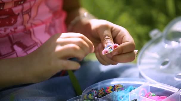 Menina criança senta-se na grama, tricota uma pulseira colorida de elásticos close-up — Vídeo de Stock