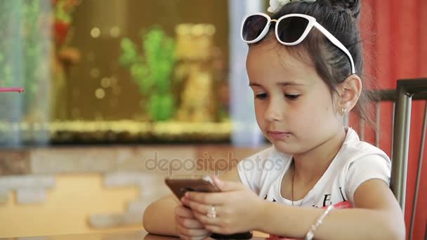 Little girl brunette sitting in cafe playing on smartphone in game with serious face degeneration — Stock Video