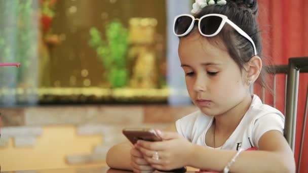 Little girl child brunette latina sitting in cafe, playing on smartphone in game, indignant, unhappy — Stock Video