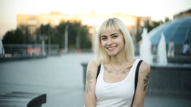 Mujer rubia joven sentada en un banco mirando a la cámara y retrato sonriente al aire libre — Vídeos de Stock