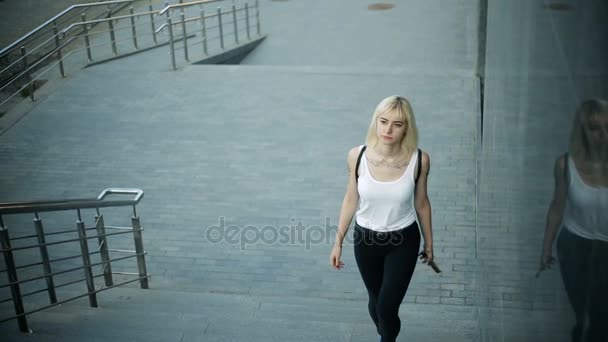 Die junge blonde Frau steigt die Treppe in der Stadt hinauf. bewegt sich auf die Kamera zu und geht vorbei. — Stockvideo