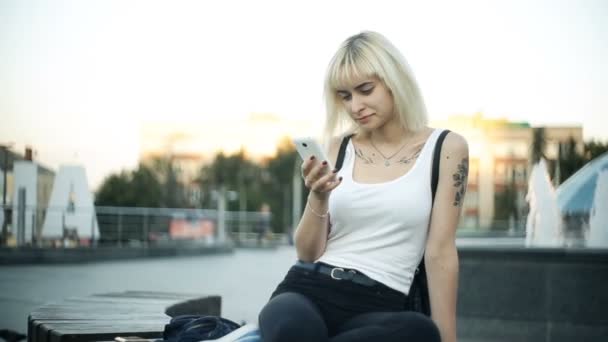 Mujer rubia joven se sienta en un banco utiliza el resplandor de teléfono inteligente de la luz del atardecer reflejado en el vidrio — Vídeo de stock