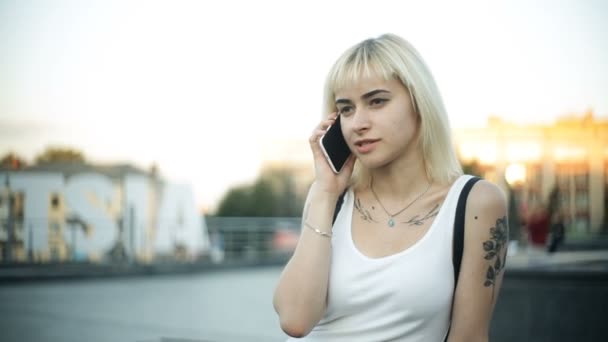 Junge blonde Frau sitzt am Telefon im grellen Licht des Sonnenuntergangs, das von Glas reflektiert wird — Stockvideo