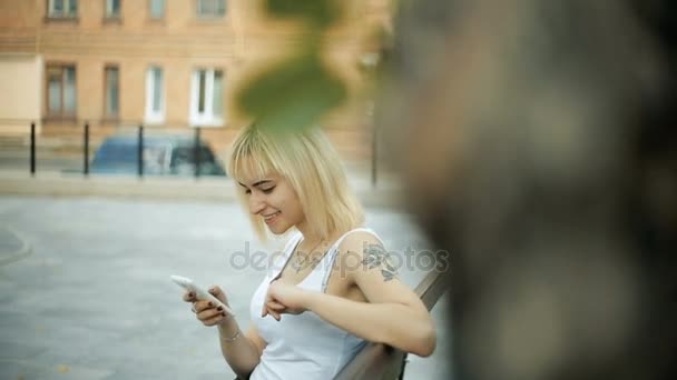 Junge blonde Frau sitzt auf einer Bank im Park nutzt ein Smartphone Internet-Text schaut sich ruhig um — Stockvideo