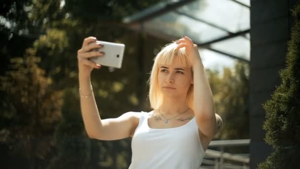 Young blonde woman looks at reflection on a smartphone straightens her hair uses the phone as mirror — Stock Video