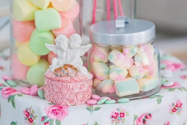 Vigselringar i smyckeskrin med änglar på en bakgrund av dekorativa glasburkar med godis marshmallows — Stockfoto
