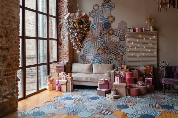 Morning interior with inverted Christmas tree suspended from the ceiling and big window. Loft interior. Beige sofa and a lot of gifts in pink colors. Hexagon tile mosaic on the wall and the floor.