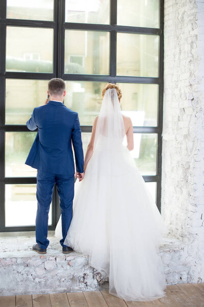 Beautiful newlyweds stand near the window in the gray studio. The bride and the pretty bride gently embrace and look into the distance. Stylish wedding in the interior