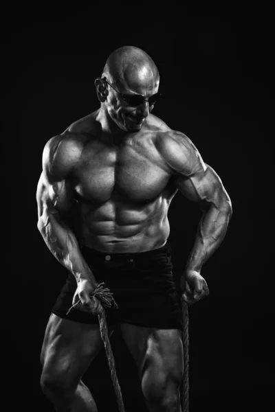 Black and white portrait of male bodybuilder with perfect six pack, posing with a rope and sunglasses — Stock Photo, Image