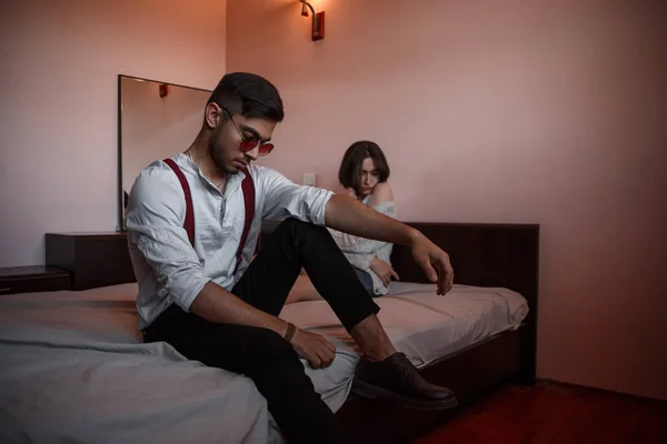 Een jonge stijlvolle man in bril zit aan de rand van het bed met — Stockfoto
