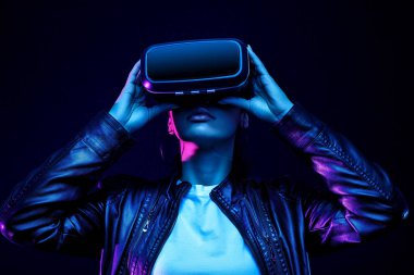 Siyah arka planda izole edilmiş, neon ışıklarla aydınlatılmış, sanal gerçeklik kulaklığıyla 360 derecelik video izleyen, VR gözlüklü Afrikalı Amerikalı kız.