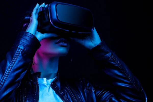 VR gözlüklü Afro-Amerikalı genç bir kadın 360 derecelik video izliyor. Sanal gerçeklik kulaklığı, neon ışıklı siyah bir arka planda izole edilmiş.