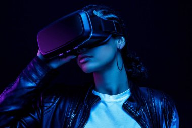 VR gözlüklü Afro-Amerikalı genç bir kadın 360 derecelik video izliyor. Sanal gerçeklik kulaklığı, neon ışıklı siyah bir arka planda izole edilmiş.