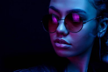 Güneş gözlüklü Afrikalı Amerikalı genç bir kadının portresi neon ışıklı siyah bir arka planda izole edilmiş.