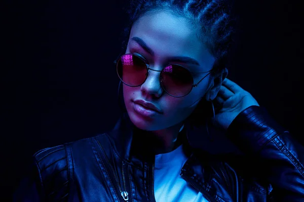 Ritratto di una giovane donna afroamericana in occhiali da sole isolata su sfondo nero in luce al neon — Foto Stock