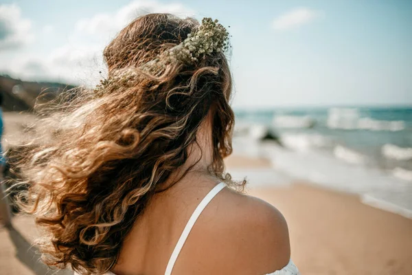 a bride in a white wedding dress on the beach with a wreath of flowers on her head. view from the back