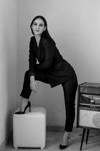 A beautiful Caucasian young woman in a black pantsuit and black sandals stands next to a vintage record player. black and white photo — ストック写真