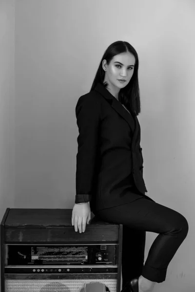 A beautiful Caucasian young woman in a black pantsuit and black sandals poses next to a vintage record player with a gramophone record in her hands. black and white image — ストック写真