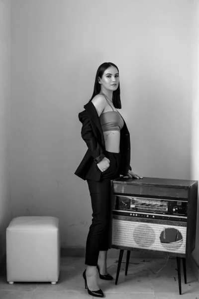 A beautiful Caucasian young woman in a black pantsuit and black sandals stands next to a vintage record player. black and white photo — ストック写真