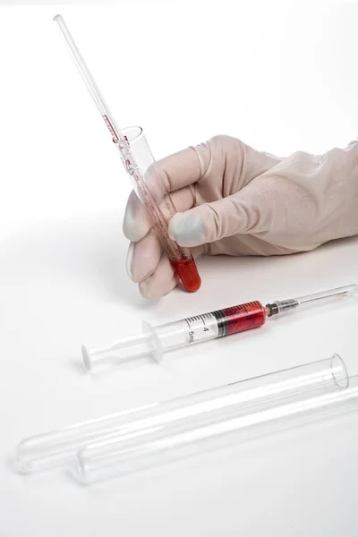 La mano enguantada de un asistente de laboratorio sostiene un tubo de ensayo con una muestra de sangre sobre un fondo blanco. también en la foto hay una jeringa con una muestra de sangre — Foto de Stock