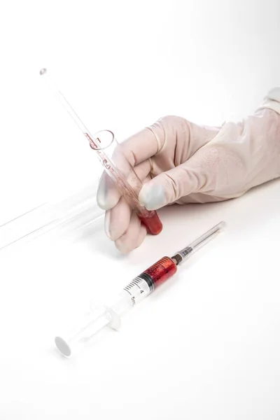 La mano enguantada de un asistente de laboratorio sostiene un tubo de ensayo con una muestra de sangre sobre un fondo blanco. también en la foto hay una jeringa con una muestra de sangre — Foto de Stock