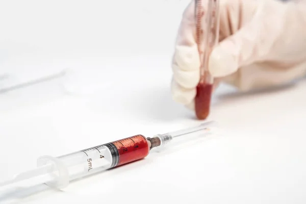 The gloved hand of a laboratory assistant holds a test tube with a blood sample on a white background. also in the photo is a syringe with a blood sample — Stock Photo, Image