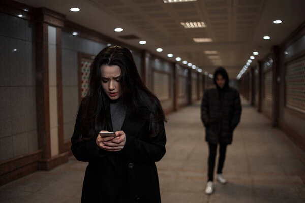 a young beautiful woman walks through an underground passage at night, followed by a man in dark clothing with a hood on his head. a woman tries to dial the number of the rescue service on the phone