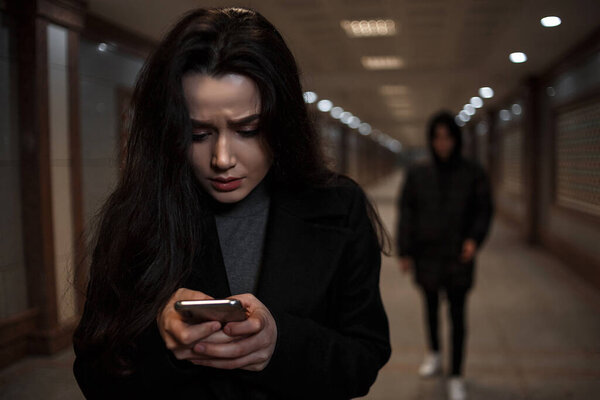 a young beautiful woman walks through an underground passage at night, followed by a man in dark clothing with a hood on his head. a woman tries to dial the number of the rescue service on the phone
