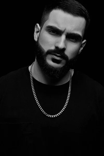 close-up of a dramatic portrait of a young serious guy, musician, singer, rapper with a beard in black clothes on a black isolated background. black and white photography.