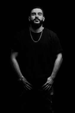 Genç, ciddi bir adamın dramatik portresini yakın plan çekiyoruz. Müzisyen, şarkıcı, siyah elbiseli, siyah izole bir arka planda sakallı bir rapçi. siyah ve beyaz fotoğrafçılık.