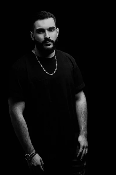 close-up of a dramatic portrait of a young serious guy, musician, singer, rapper with a beard in black clothes on a black isolated background. black and white photography.