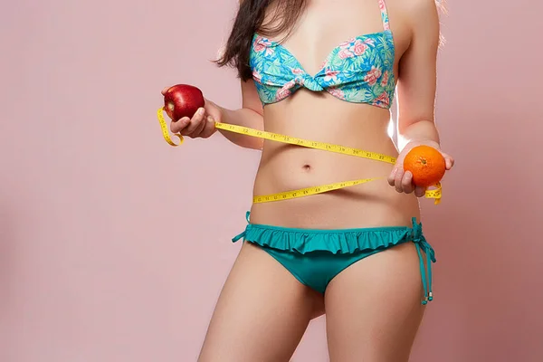 水着姿のアジア系の若い美少女 片手にオレンジ 左手にアップル 手に測定テープを持っています ピンク色の孤立した背景で — ストック写真