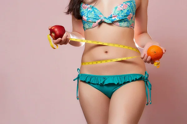 水着姿のアジア系の若い美少女 片手にオレンジ 左手にアップル 手に測定テープを持っています ピンク色の孤立した背景で — ストック写真