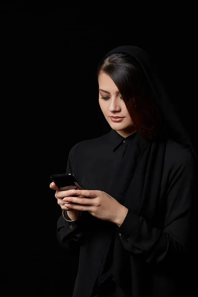 a young beautiful Muslim woman in black clothes uses a mobile phone.smiling, texting, talking. on a black isolated background.