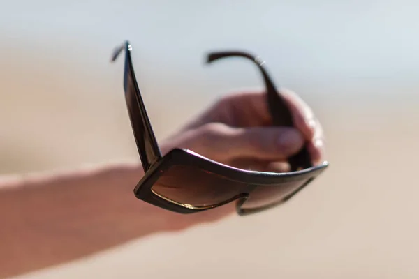 A man's hand holds large brown sunglasses. Blurred background of a sunny beach. — Stock Photo, Image