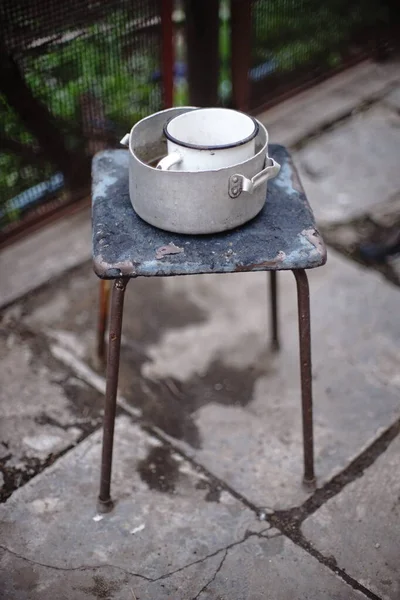 Старий посуд на ретро табуреті. Алюмінієва сковорода та емальований кухоль на стільці. Сільський двір . — стокове фото