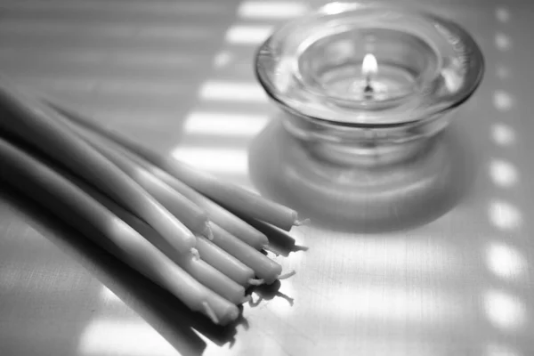 Bunch of new long candles and one low candle burns in a glass round candlestick, bw photo — Stock Photo, Image